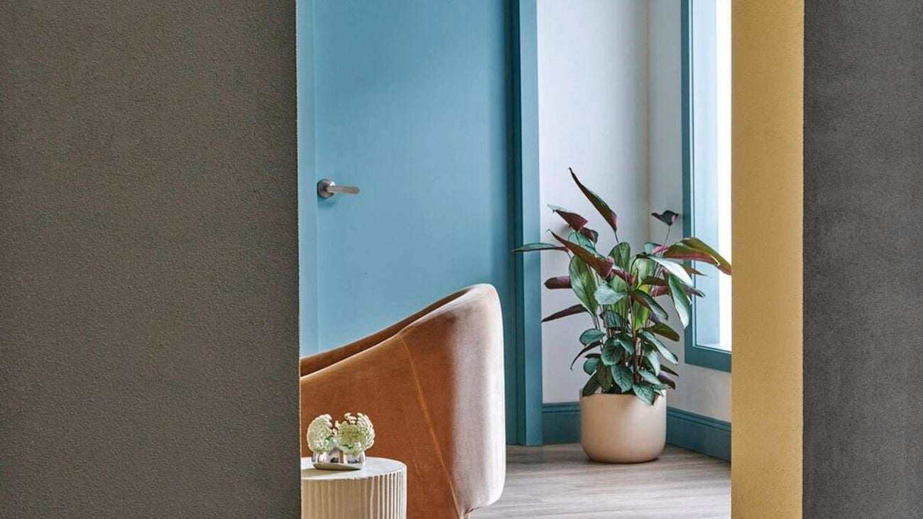 Grey wall with arch doorway, opening out into a painted blue room with pot plants and a brown sofa chair.