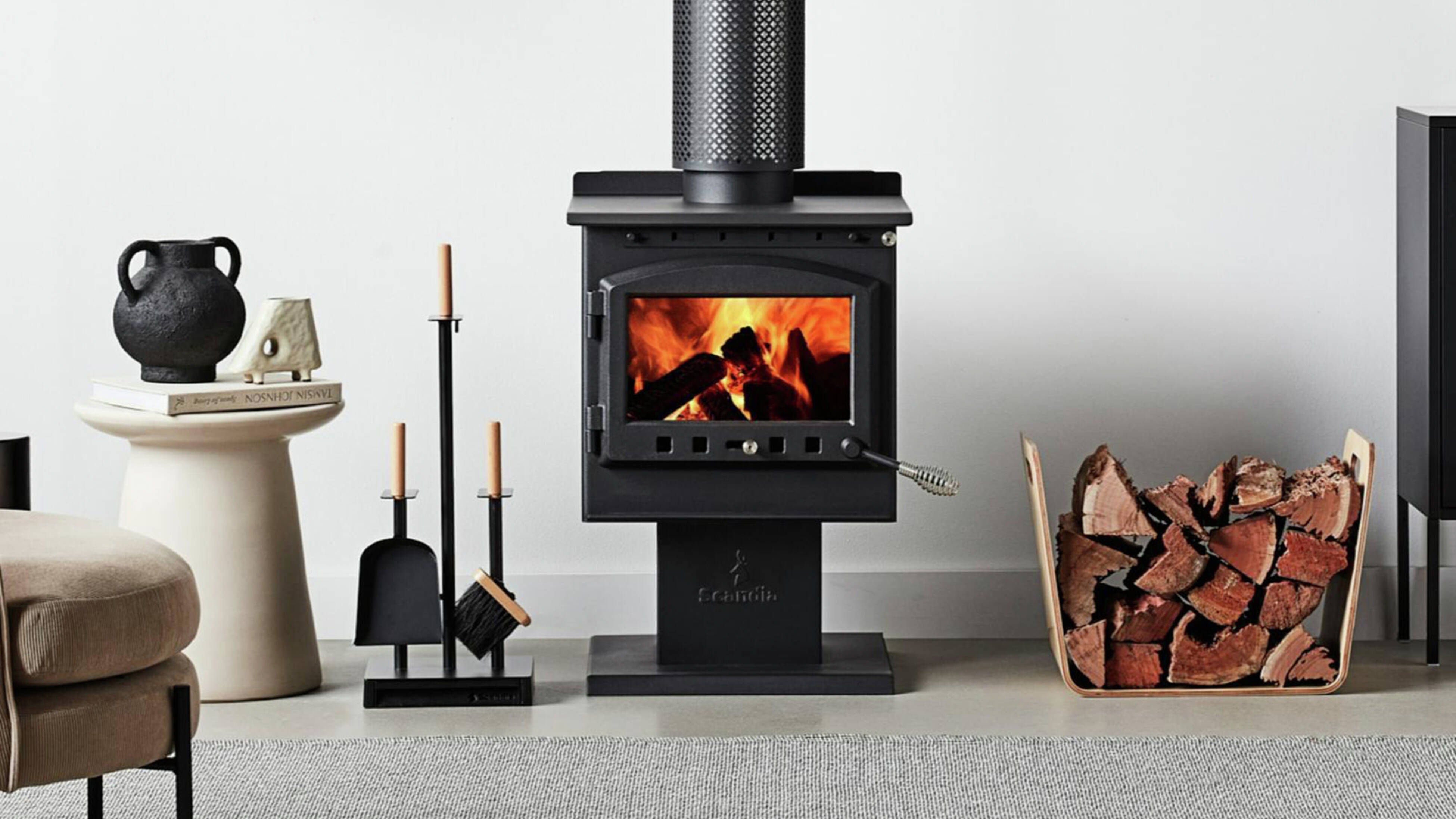 Freestanding lit black wood‑burning stove with a flue pipe, fireplace tools to the left and stacked logs to the right
