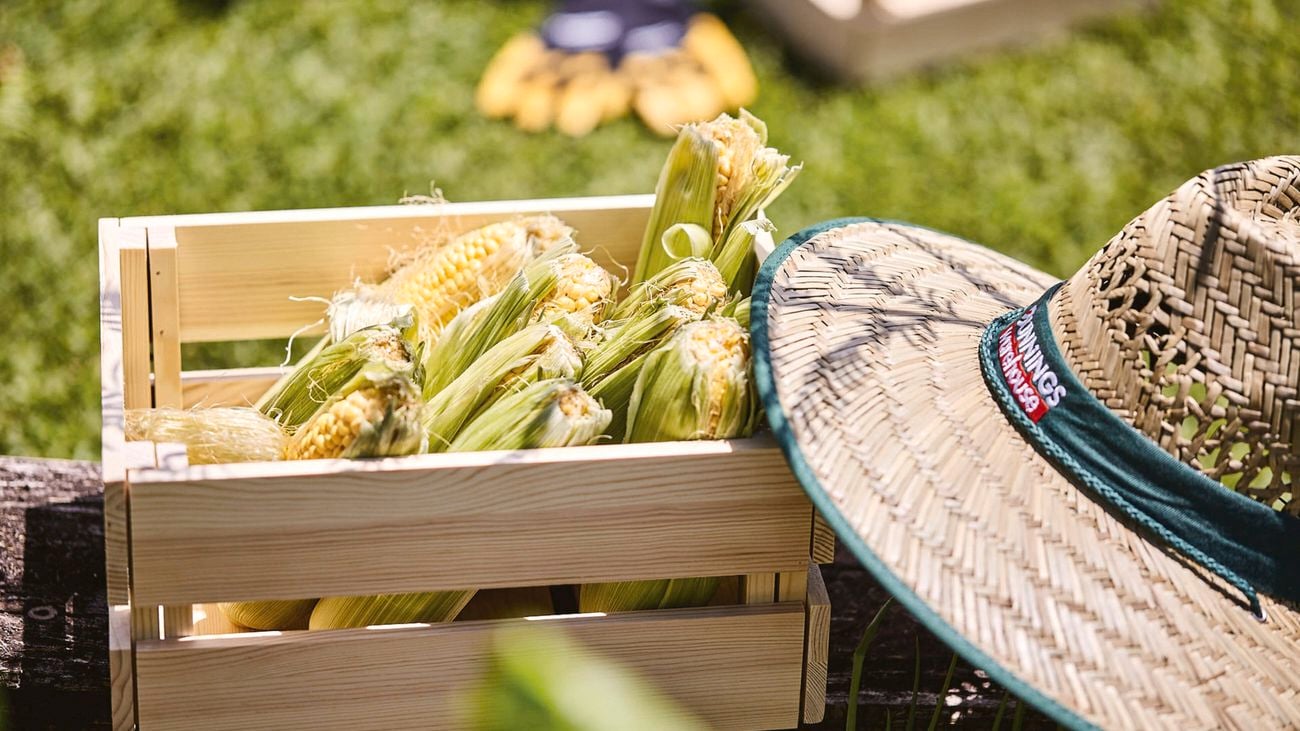 Corn in its husk, sitting in a wooden crate on the grass, beside a Bunnings straw hat.