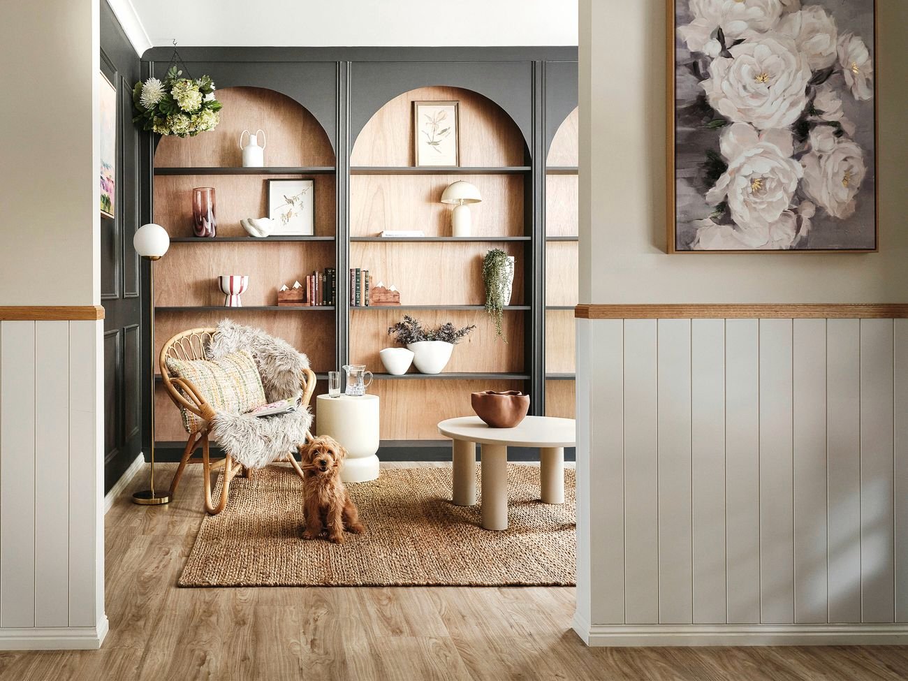 Modern living room with dark built-in shelving featuring arched niches, styled with books, ceramics and plants, alongside a rattan chair, side tables and a small dog on a woven rug.
