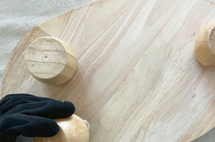 A gloved hand attaching a bun foot to a wooden chopping board with glue