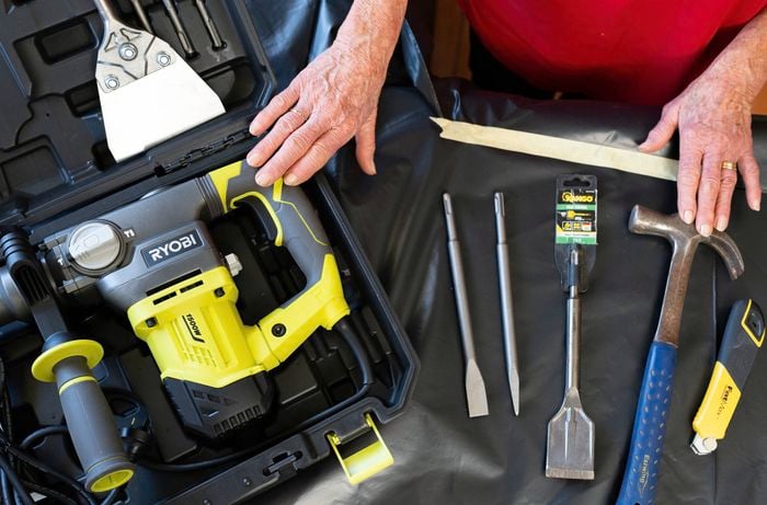 A selection of tools, including rotary hammer drill with chisel bits, a hammer, and a utility knife, are displayed on a table. 