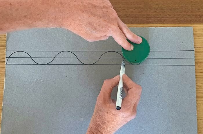 A pair of hands using a spray can lid and marker to make a scallop template on cardboard