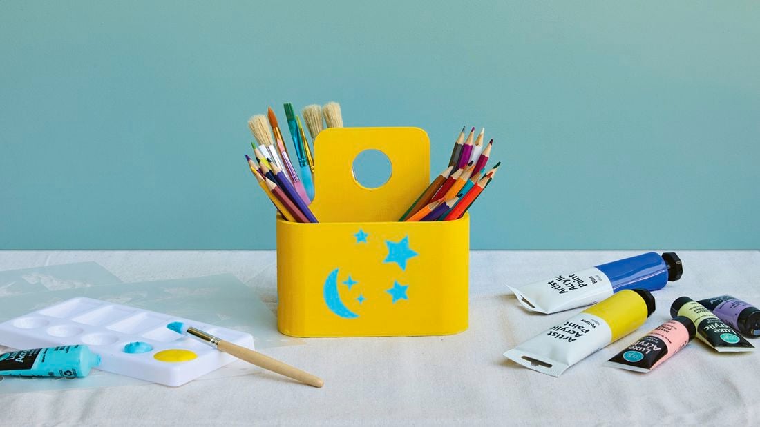 A bright yellow wooden art caddy decorated with blue stencilled stars and a moon, filled with coloured pencils and paintbrushes, surrounded by paint tubes and a paint palette.