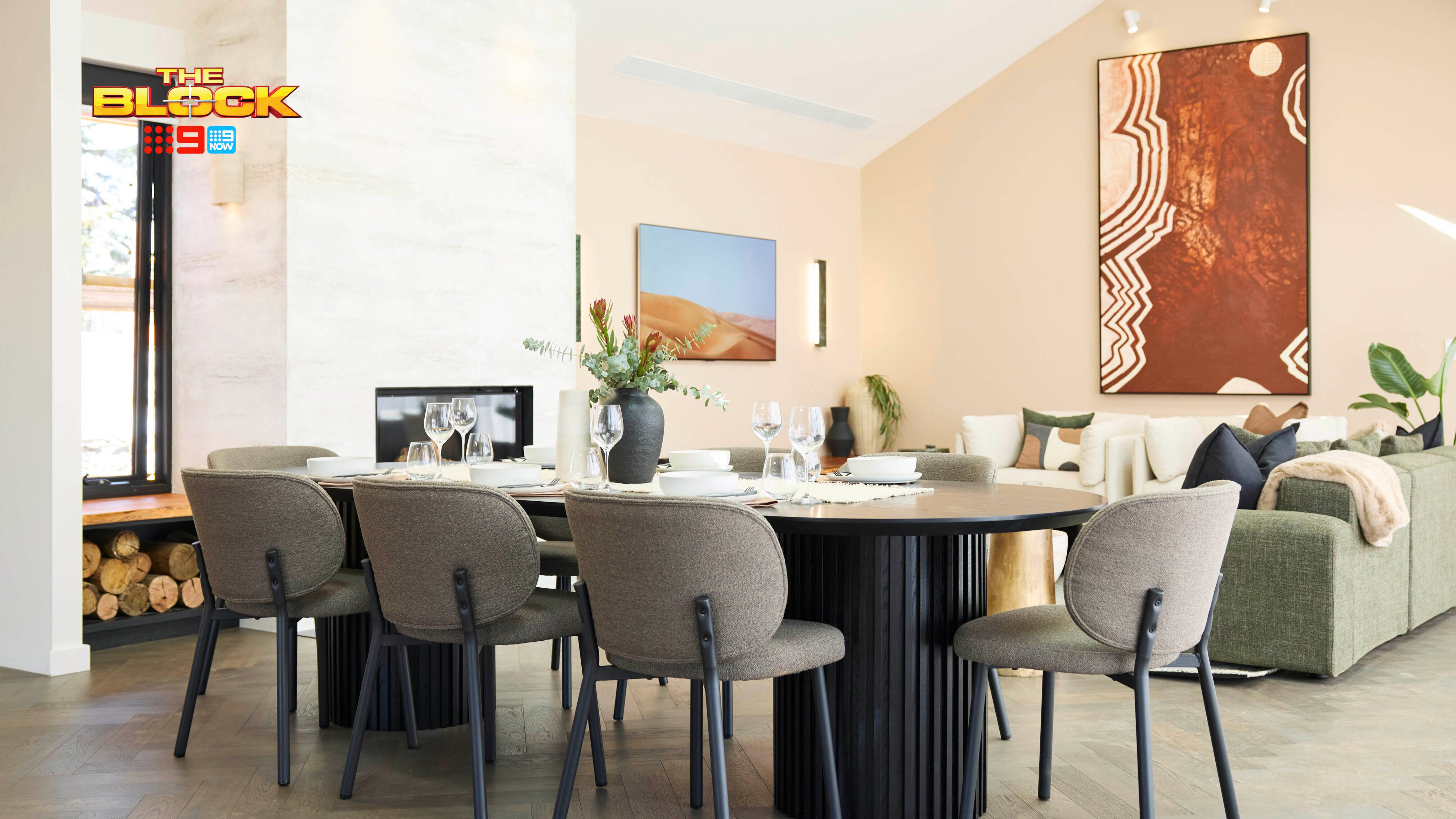A modern dining area with a round table surrounded by six upholstered chairs. The table is set with plates, glasses, cutlery, and a floral centerpiece. In the background, a living room features a green sofa with cushions and two pieces of wall art.