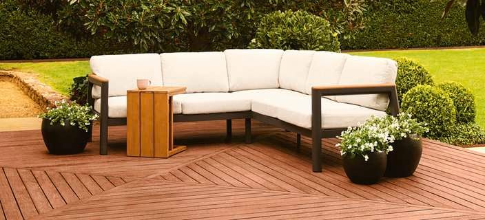 Outdoor corner lounge with a wooden side table on a timber deck, surrounded by green foliage.