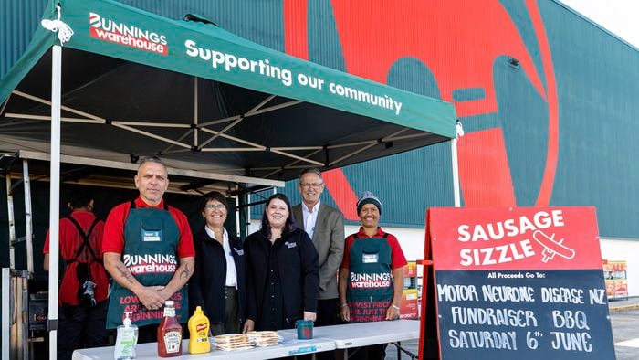 Six adults stand behind a sausage sizzle stall outside a Bunnings Warehouse store. The group is under a green canopy printed with “Bunnings Warehouse” and “Supporting our community,” with a barbecue setup behind them and a table in front holding bread, sauce bottles, and a small container. A large red sign to the right reads “SAUSAGE SIZZLE,” and a chalkboard sign says “All proceeds go to: Motor Neurone Disease NZ, Fundraiser BBQ, Saturday 6th June.” The background shows the store’s exterior with large red and green wall graphics.
