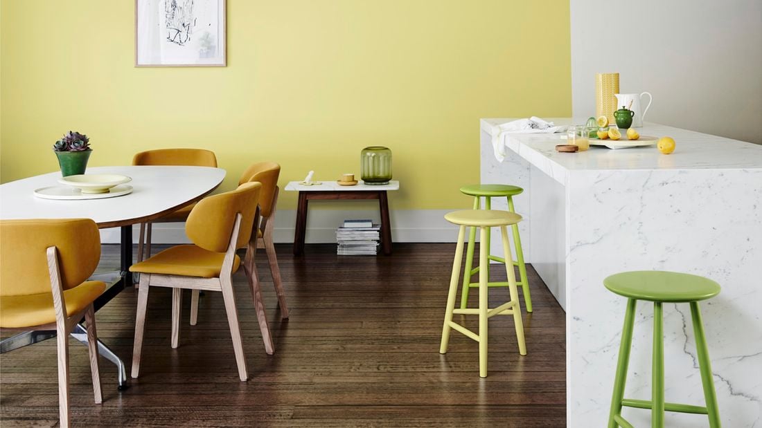 A kitchen with timber floors, marble bench, yellow chairs and stools and a yellow feature wall