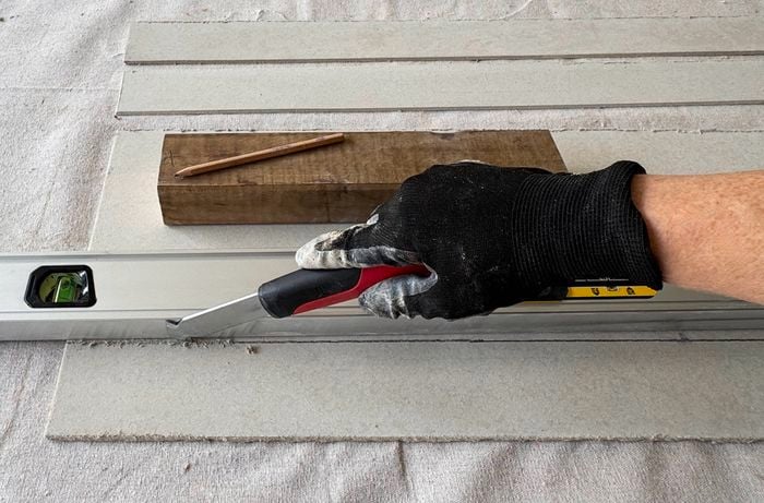 A gloved hand scores a fibre‑cement sheet with a utility knife guided by a spirit level.