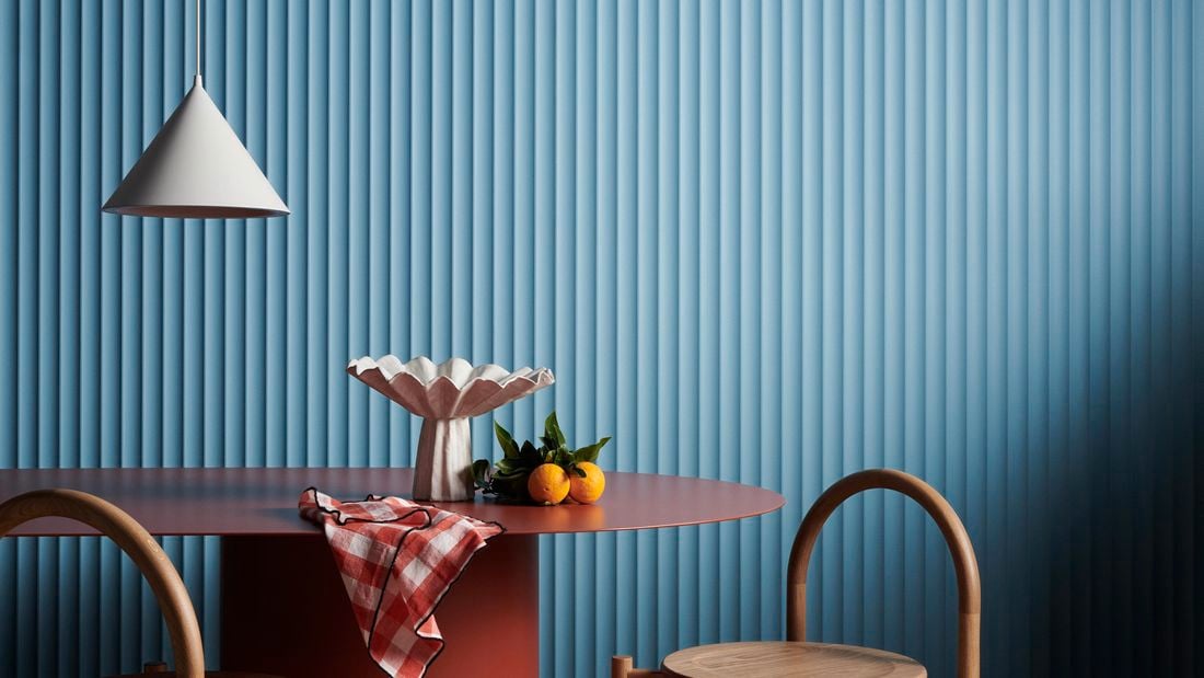A hanging lamp, table and two chair in front of a blue Laminex wall.