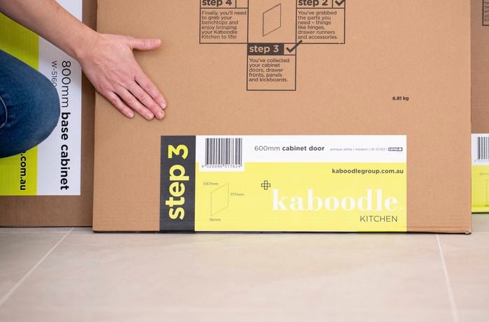 A person is handling a flat-pack kitchen cabinet box, part of a DIY kitchen renovation setup.