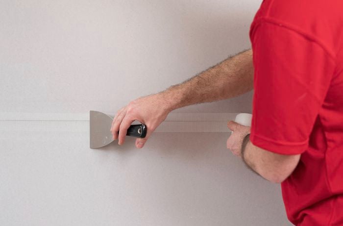 A person in a red T-shirt presses jointing tape with one hand while holding a trowel in the other