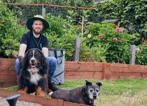 Ash Burke seated in a garden with two dogs, one large and one medium-sized, and a garden bag in the background. A garden with raised wooden beds filled with flowering plants, a gravel path and a wooden gate in the foreground.