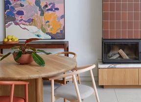 Modern dining area with a round wooden table and chairs, a white pendant light, a colourful abstract painting, and a built‑in fireplace clad in terracotta tiles with a black flue and timber base cabinet.