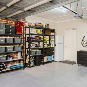 Organized garage with shelves holding plastic bins and tools, a black cabinet on the left, and a white refrigerator against the back wall. The open garage door reveals ceiling tracks and opener mechanism. 