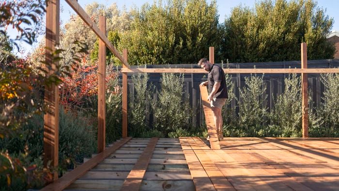 A person installs wooden deck boards on a raised outdoor structure, standing on partially laid timber decking inside a timber frame, with garden plants and a fence in the background.