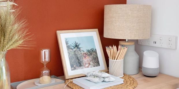 Hallway table with orange feature wall and accessible power points. The table has various objects on it such as an hourglass, a picture of a girl at the beach, a lamp, a tin of pencils and a Google home.