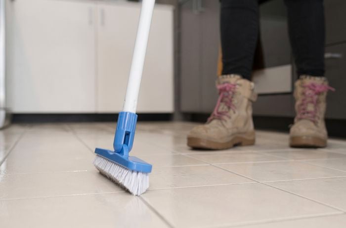 A person is cleaning grout lines with a long-handled grout brush.