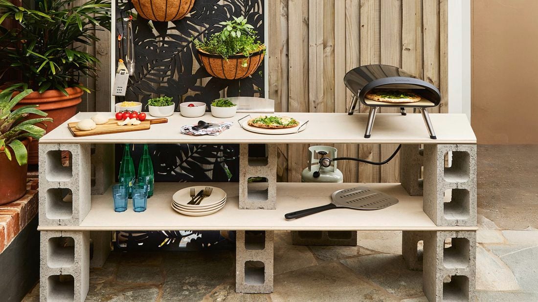 A besser block outdoor kitchen bench space with pizza oven, utensils, bowls of herbs and a timber chopping board with vegetables on it.