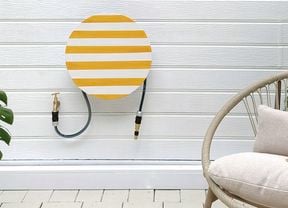 A garden patio with a large potted Monstera plant, a striped hose hanger cover on a white wall, and a woven outdoor chair
