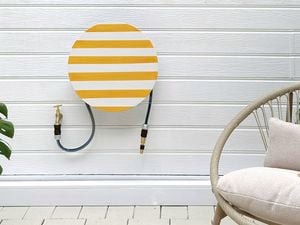 A garden patio with a large potted Monstera plant, a striped hose hanger cover on a white wall, and a woven outdoor chair