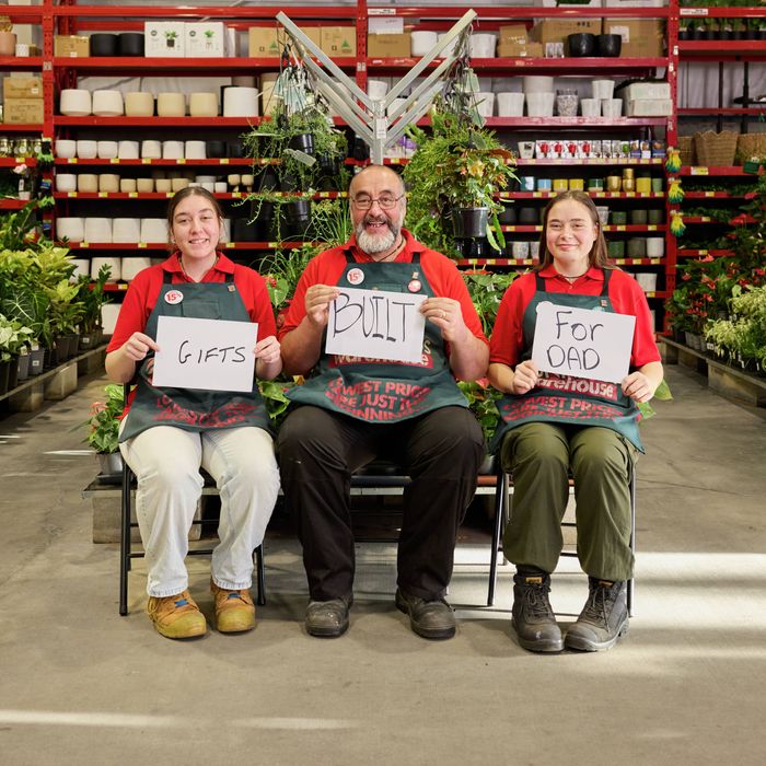Three Bunnings team members seated in the plant section of a store, surrounded by shelving stocked with pots and gardening products. Each person holds a handwritten sign that together reads “GIFTS BUILT FOR DAD.” Hanging plants and rows of potted greenery are visible behind them.