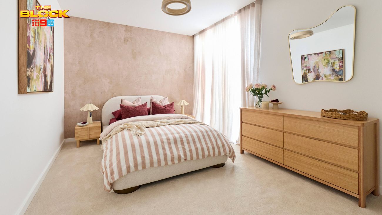 Contemporary bedroom featuring striped neutral bedding, a curved bedhead, timber dresser, statement mirror and soft pink feature wall.