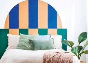 Bedroom with a painted geometric half-arch headboard in blue, peach and green, white bedding with layered cushions, bedside table and indoor plant.