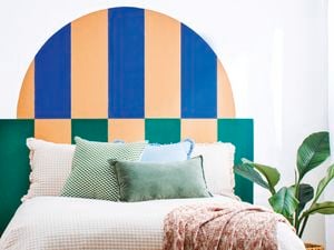 Bedroom with a painted geometric half-arch headboard in blue, peach and green, white bedding with layered cushions, bedside table and indoor plant.