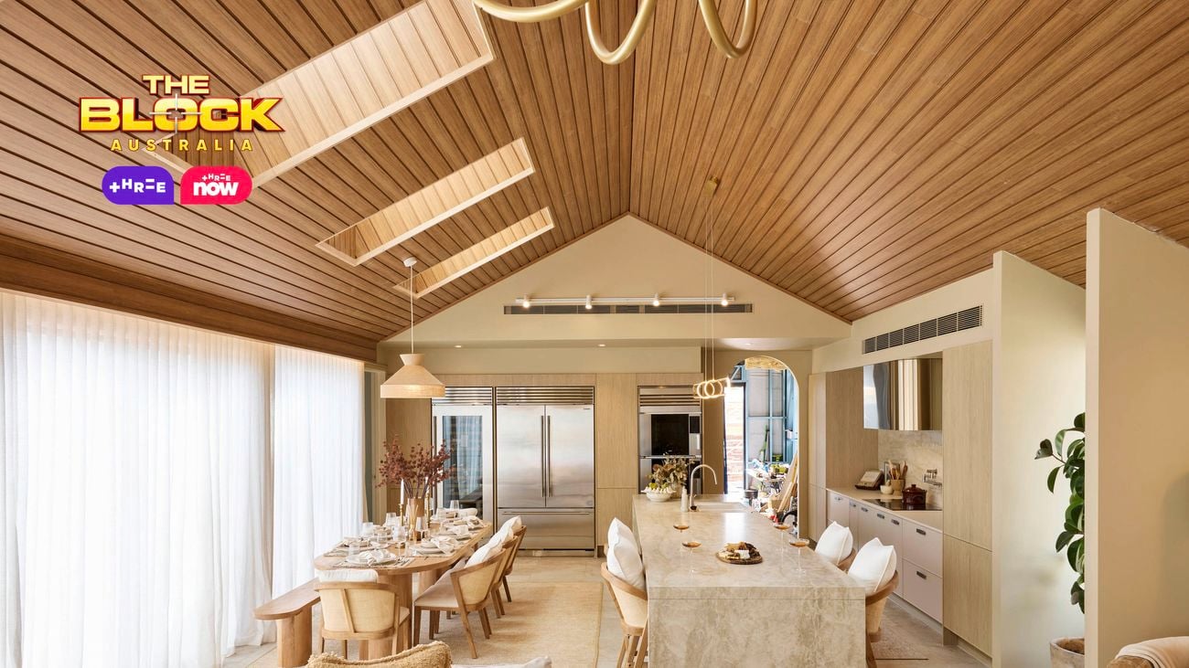 Open-plan kitchen and dining space from The Block, featuring a vaulted timber ceiling, marble island with bar stools, large fridge, neutral-toned dining area, and floor-to-ceiling curtains.