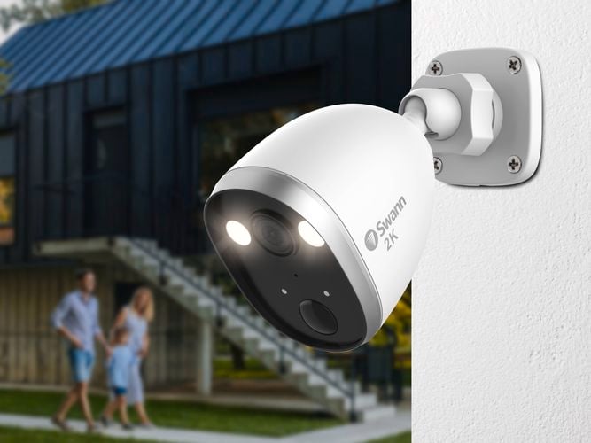 Swann 2K security camera mounted on a white wall, with a modern building, external staircase, and three people walking on grass in the background.