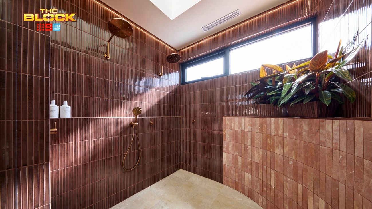 Modern shower with maroon tiled walls, dual gold rain showerheads, light tiled floor, and a planter near a window. 