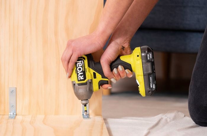 A person securing an angle bracket to a timber shelf with a drill
