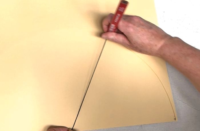 Hands holding a string at the corner of a cream piece of card, and using a pencil to draw an arc on the paper.