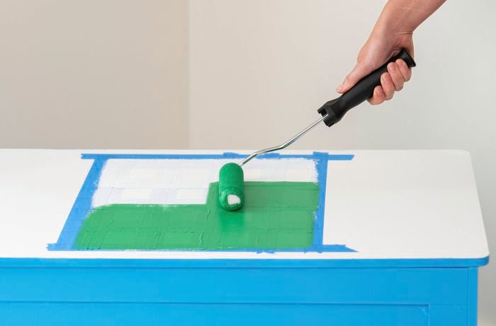 A person is painting a green square on the toy box lid with a paint roller using painter’s tape.