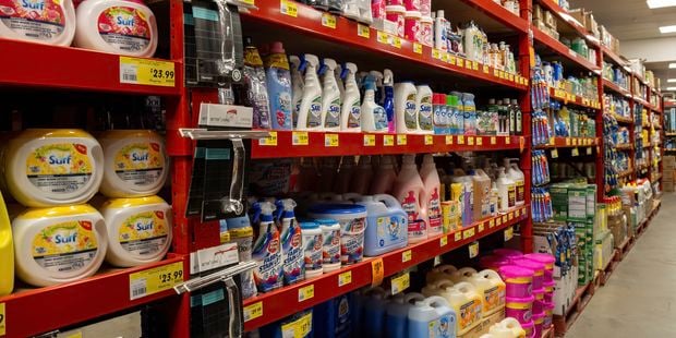 Aisle of cleaning and laundry products at a Bunnings store, with bright orange shelving stocked with detergents, stain removers, fabric softeners, disinfectants, spray cleaners and household supplies. Yellow price labels line the shelves, and the aisle extends into the distance under fluorescent lighting.