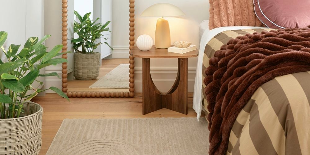 A styled bedroom corner with a bed dressed in warm-toned textiles, a wooden bedside table with a lamp, a beaded-frame floor mirror, and potted plants on timber flooring.