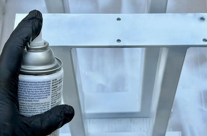 A gloved hand spraying metal table legs with white spray paint on top of a drop sheet.