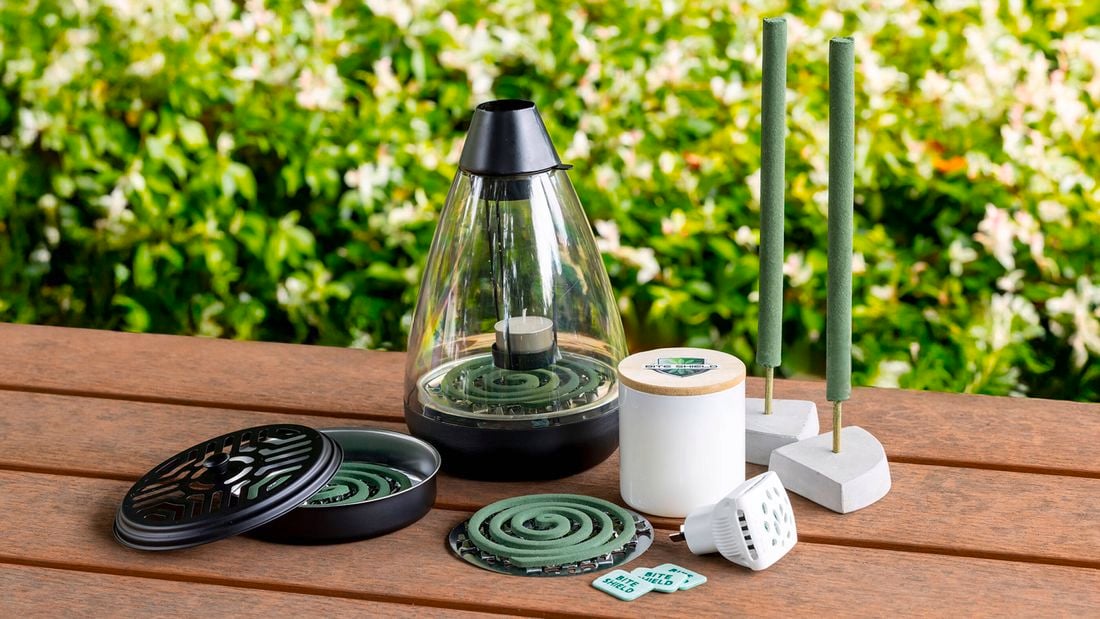 Close up of a timber outdoor table with greenery behind with natural mosquito repellent, mosquito coils plus a candle and plug-in diffuser on top