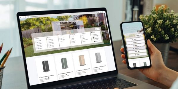 A person holding a mobile phone ahd sitting in front of a laptop with the Select A Shed app.