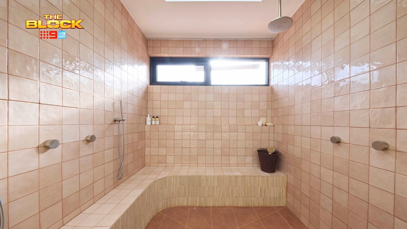 Spacious shower room with pink tiled walls, brown tiled floor, built-in bench, multiple showerheads, and natural light from a high window. 