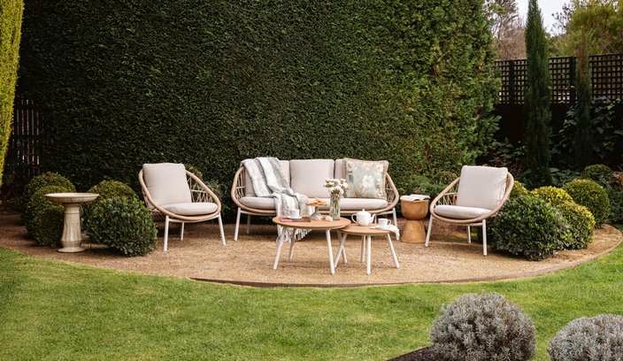 Cane outdoor setting outside on a paved area with hedging behind it.