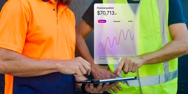 Two workers wearing high‑visibility safety clothing stand together outdoors, using a tablet. A transparent digital overlay shows a financial dashboard with a line graph and the text “Financial position $70,713,” indicating income, expenses, and profit over time.