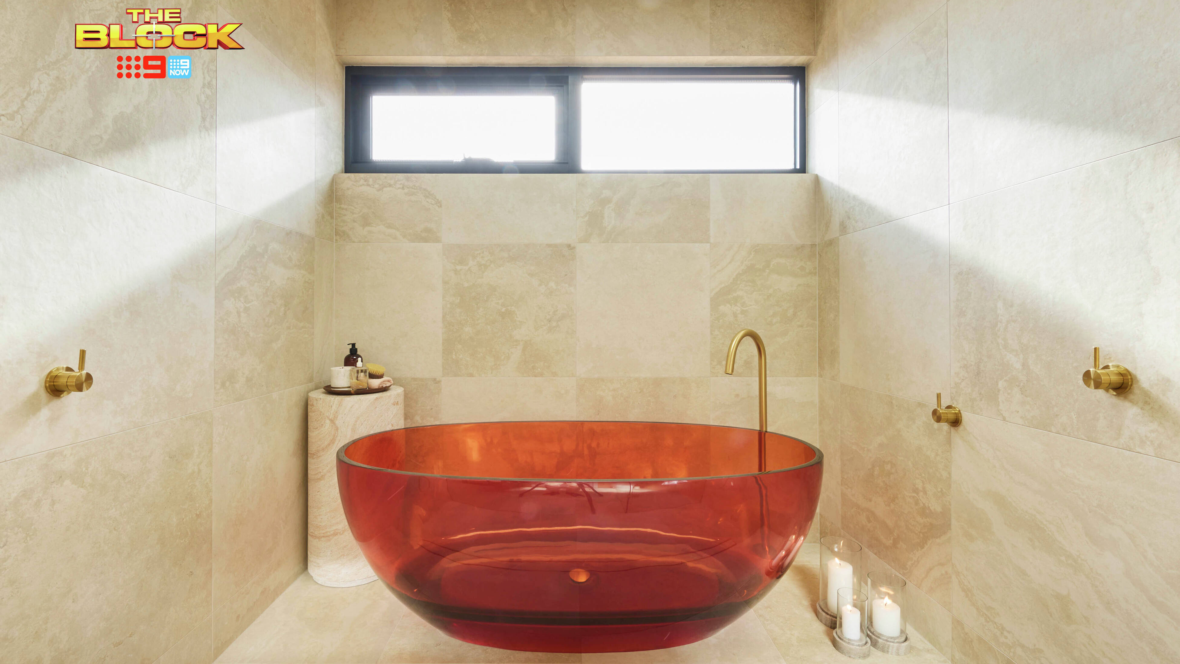 Modern bathroom with translucent amber tub, gold faucet, beige tiles, bath products on a stand, candles on the floor, and natural light from a window. 