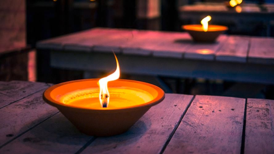 "Close-up of a lit candle in an orange bowl on a wooden table, with another similar candle in the background in a dimly lit outdoor or semi-outdoor setting.