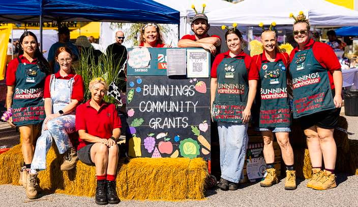 Group of Bunnings Team Members standing and sitting around a Bunnings Community Grants sign at an outdoor community event with market tents in the background.