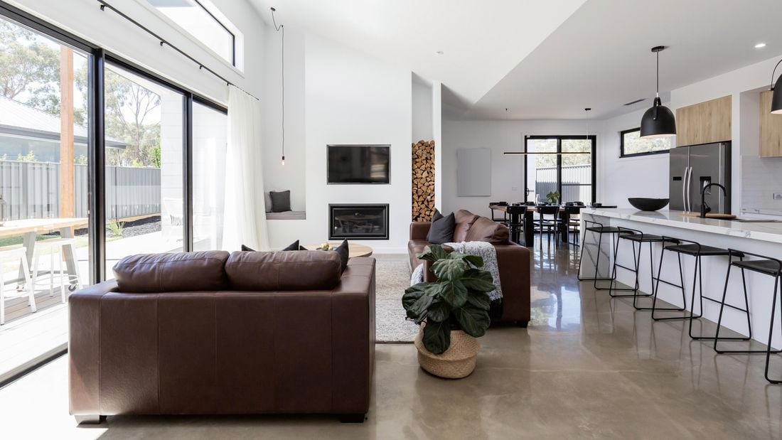 Modern living room with brown leather couches, concrete floors and a fireplace next to a tall stack of wood logs.