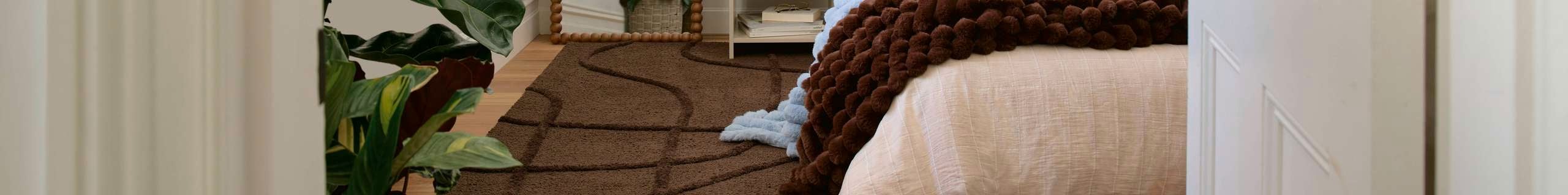 View into a bright, minimal bedroom with a low bed dressed in neutral bedding and brown throw, a brown rug, a tall floor mirror, a leafy indoor plant, and a white bedside table near tall window