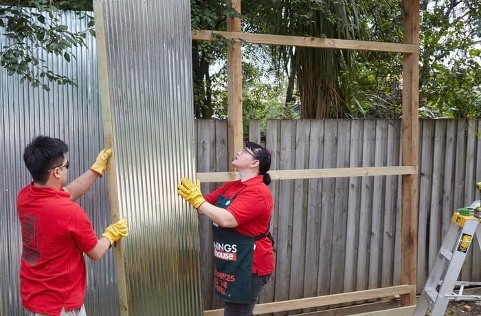 Two people wearing work gloves lifting a sheet of corrugated iron into position against fence rails