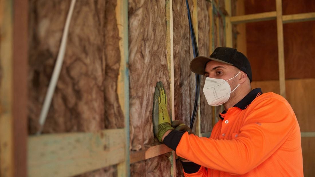 A tradesperson in an orange shirt, gloves, cap, and face mask installs fibrous insulation into a timber-framed wall cavity.]
